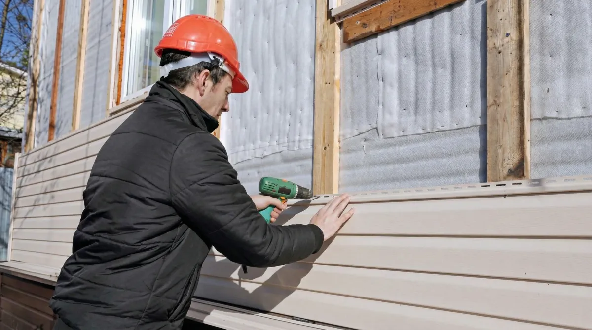 Siding replacement professional using a power drill to install new tan vinyl siding on a home exterior in Lowell, MA